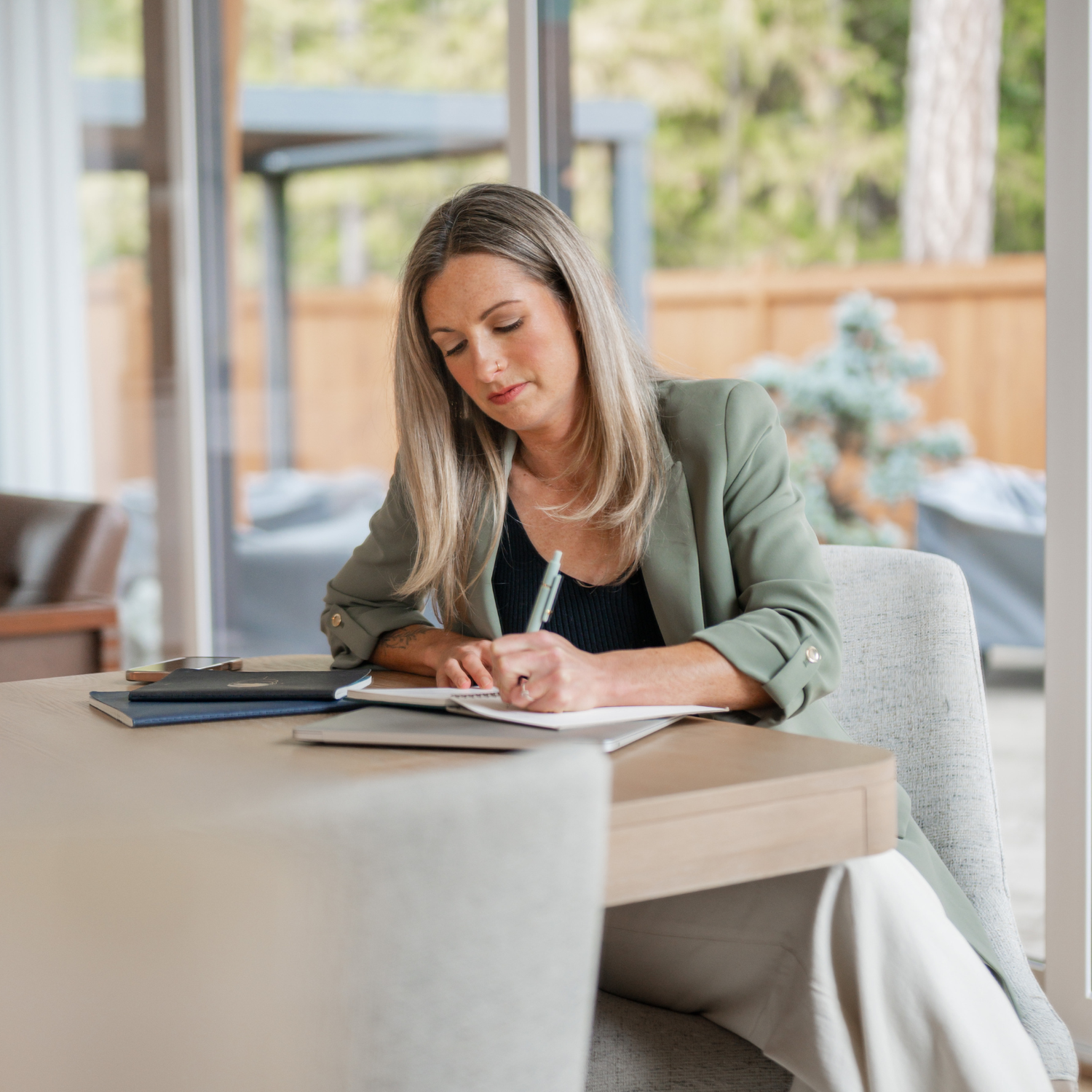Woman in a green blazer reviewing a coaching contract at a desk by a forest-view window