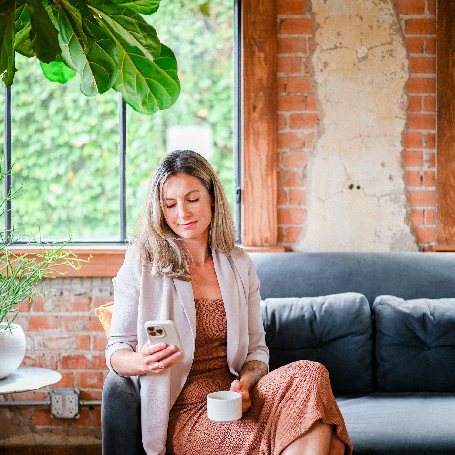 Jaime Bell, Lawyer sitting on a coach reading her phone. Wearing a brown dress and beige jacket, tree in background with brick wall